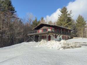 A two-story chalet with a balcony, surrounded by snow and trees.