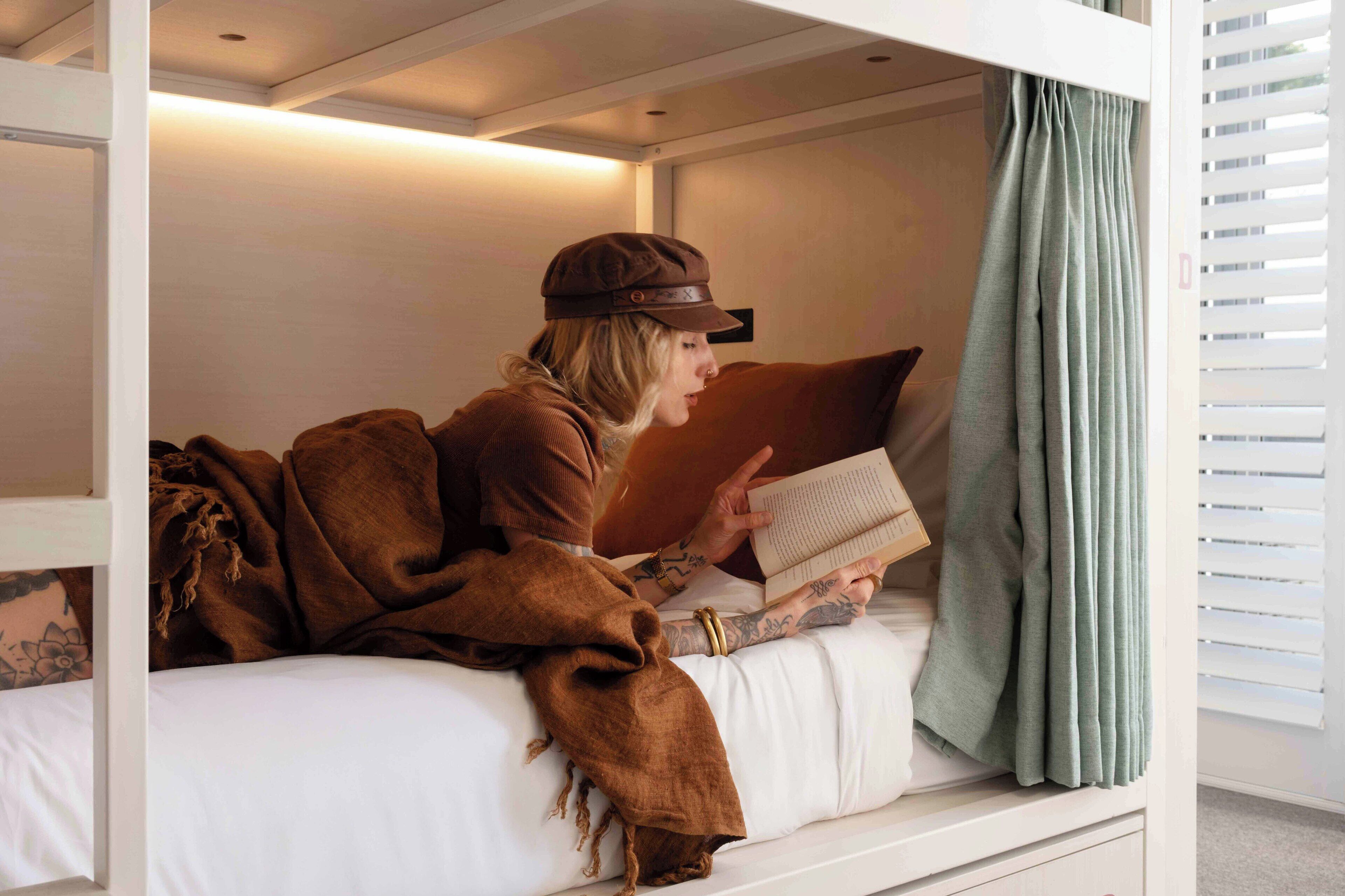 A person reading a book in a room with a bunk bed, a blanket, and a curtain.