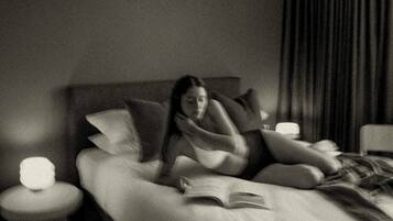 Una persona recostada en una cama con almohadas, un libro abierto y una lámpara de noche.
