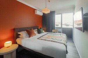 Habitación de hotel con una cama grande, una mesita de noche con lámpara, una luz en el techo y vistas al exterior a través de una puerta corredera.