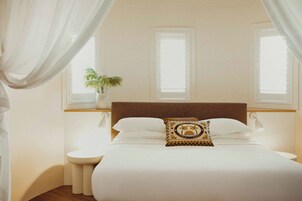Una cama bien tendida con sábanas blancas y una almohada decorativa, una planta en la mesita de noche y una ventana con persianas.