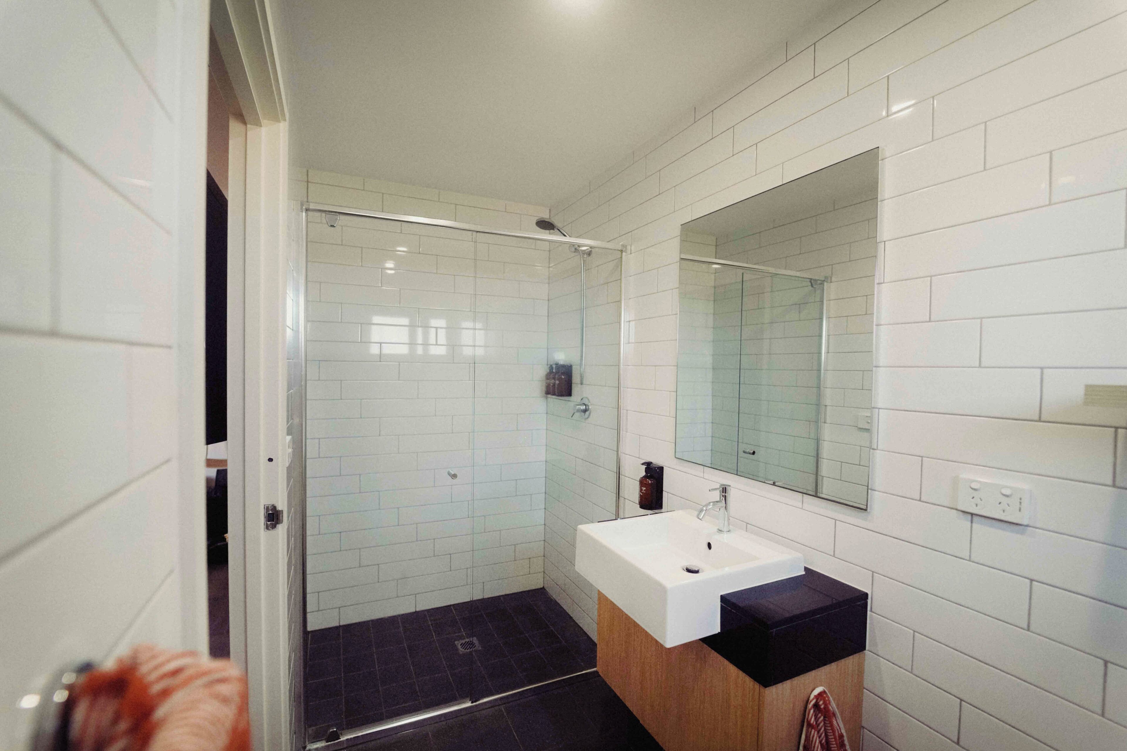 A modern bathroom with white tiles, a glass shower enclosure, a white sink, and a large mirror.