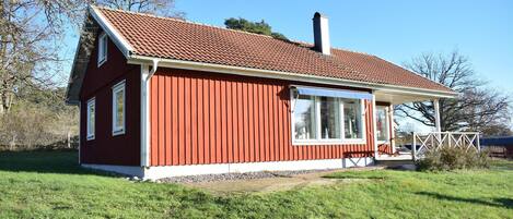 A red wooden house with a tiled roof, a chimney, and a porch with a white railing.