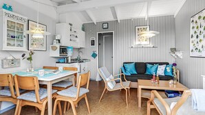 A modern kitchen with white cabinetry, a dining area with wooden chairs, and a living room with a sofa and cushions. - Amazing home in Allinge (Allinge)