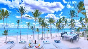 A beachside dining area with a table set for two, surrounded by lounge chairs and a view of the ocean. - Punta Cana Ocean View Penthouse - The Best Dominican Ocean View (Punta Cana)
