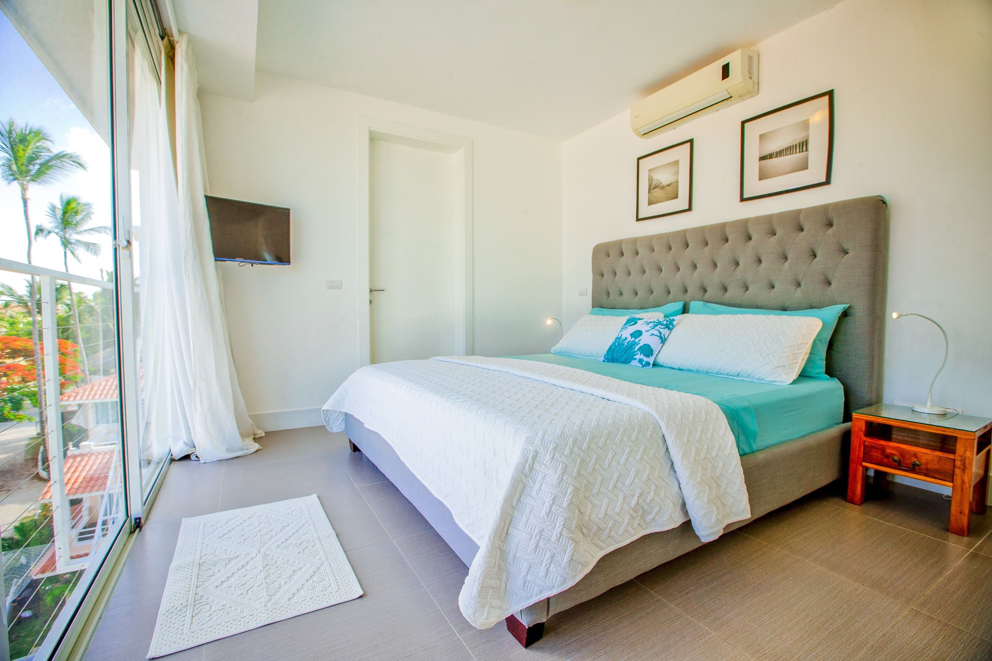 Un dormitorio con una cama grande, una mesita de noche, un televisor montado en la pared y vistas a palmeras a través de la ventana.