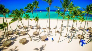 A beach with palm trees, thatched umbrellas, and beach chairs. - Punta Cana Ocean View Penthouse - The Best Dominican Ocean View (Punta Cana)
