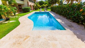 A rectangular swimming pool with a tiled deck, surrounded by lush greenery and palm trees. - Punta Cana Condo With Swimming Pool - Bavaro Condo Cocotal Sanitized (Punta Cana)