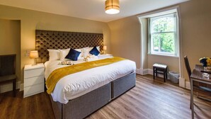 A bedroom with a tufted headboard, a bed with white linens and a yellow throw, bedside lamps, a small table with a vase, and a window with a view of greenery. - Rosedene Highland House - Emperor Apartment 7 (Inverness)
