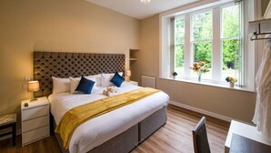 Ein Schlafzimmer mit einer gesteppten Kopfteile, einem großen Bett mit weißer Bettwäsche und blauen Kissen, einem Nachttisch mit Lampe, einem Fenster mit Blick auf Grünflächen und einer Heizung.