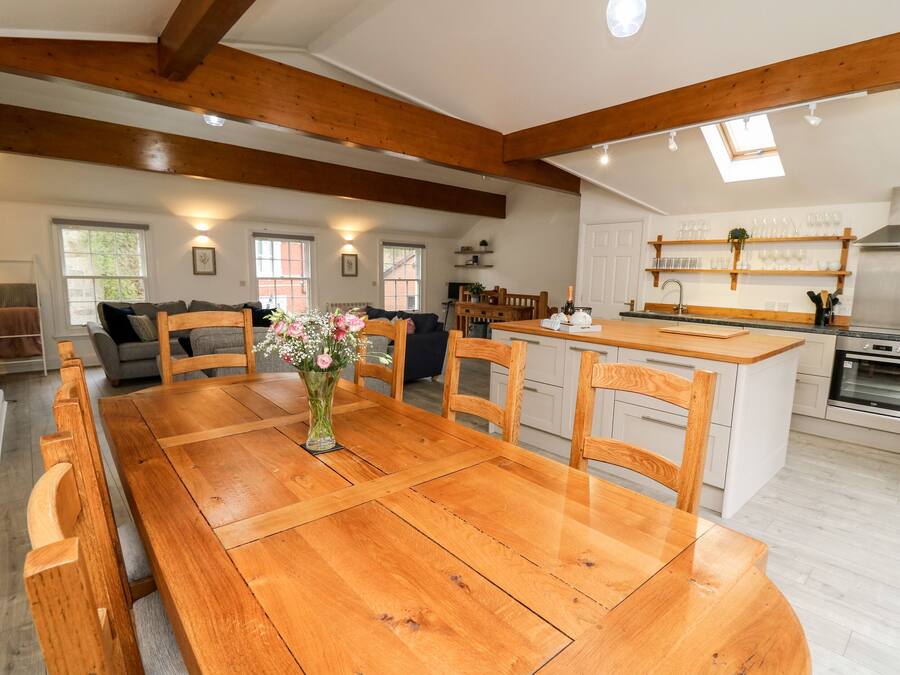 A spacious kitchen with wooden beams, a dining area with a wooden table and chairs, and a view of the living room with a sofa and a vase of flowers.