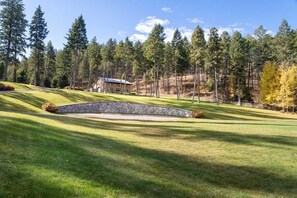Un campo da golf curato, con un bunker in pietra, una casa sullo sfondo e una fitta foresta circostante.