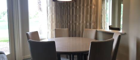 A dining area with a round table, six chairs, a hanging light fixture, and a window with curtains.