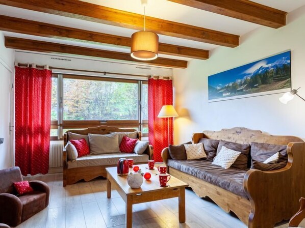 Un salon chaleureux, avec des meubles en bois, une grande fenêtre ornée de rideaux rouges, un tableau accroché au mur et une table sur laquelle sont posées des tasses rouges.
