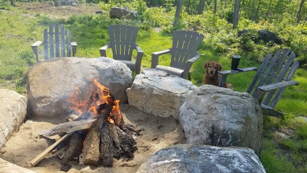 A campfire with logs and rocks, surrounded by Adirondack chairs and a dog. - Mountain View Cabin (Lovell)