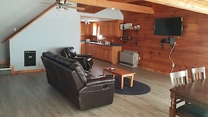 A living room with a sofa, a coffee table, and a dining table with chairs. There is a kitchen area with cabinets and a refrigerator. A ceiling fan is visible, and a television is mounted on the wall. - Mountain View Cabin (Lovell)
