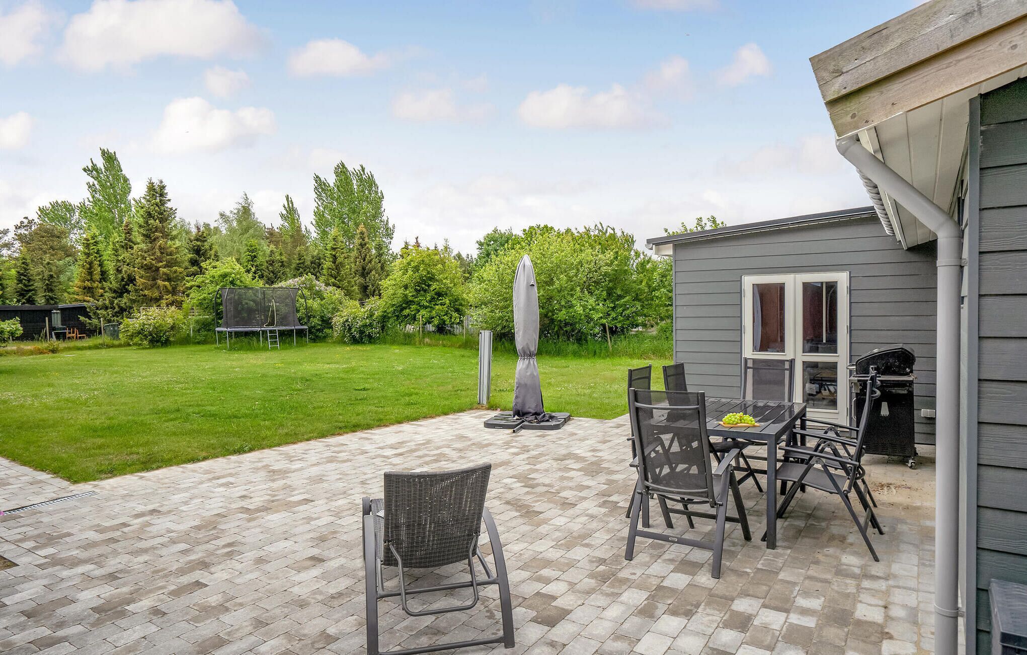 Een terras met tuinmeubilair en een barbecueplaats, omgeven door een gazon en bomen.