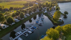 Eine Vogelperspektive zeigt einen Seeufer-Campingplatz mit Wohnmobilen und Booten, die an einem schmalen Wasserweg liegen.