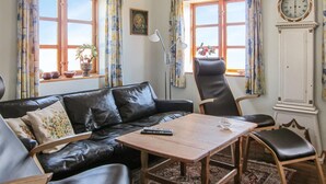 Una sala de estar con un sofá de cuero negro, una mesa de centro de madera y un sillón de cuero negro. Hay dos ventanas con cortinas, una lámpara de pie y un reloj de pared.
