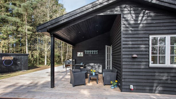 A wooden cabin with a covered patio area featuring outdoor furniture and a small table with a vase of flowers. - Amazing home in Nexø with kitchen (Nexø)