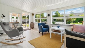 A spacious living room with a rocking chair, a blue armchair, and a sofa. There are large windows offering views of the garden and a patio area. - Gorgeous home in Nexø with WiFi (Nexø)