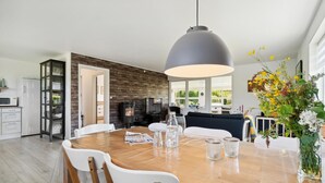 A modern kitchen with a wood-burning stove, a dining area with a wooden table and white chairs, and a large window providing natural light. - Gorgeous home in Nexø with WiFi (Nexø)