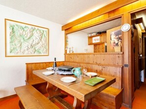 A dining area with a wooden table set for two, a green plate, wine glasses, and a bottle. A framed abstract painting hangs on the wall.