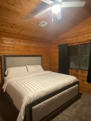 Quarto com painéis de madeira, cama, ventoinha no teto e janela com cortinas.