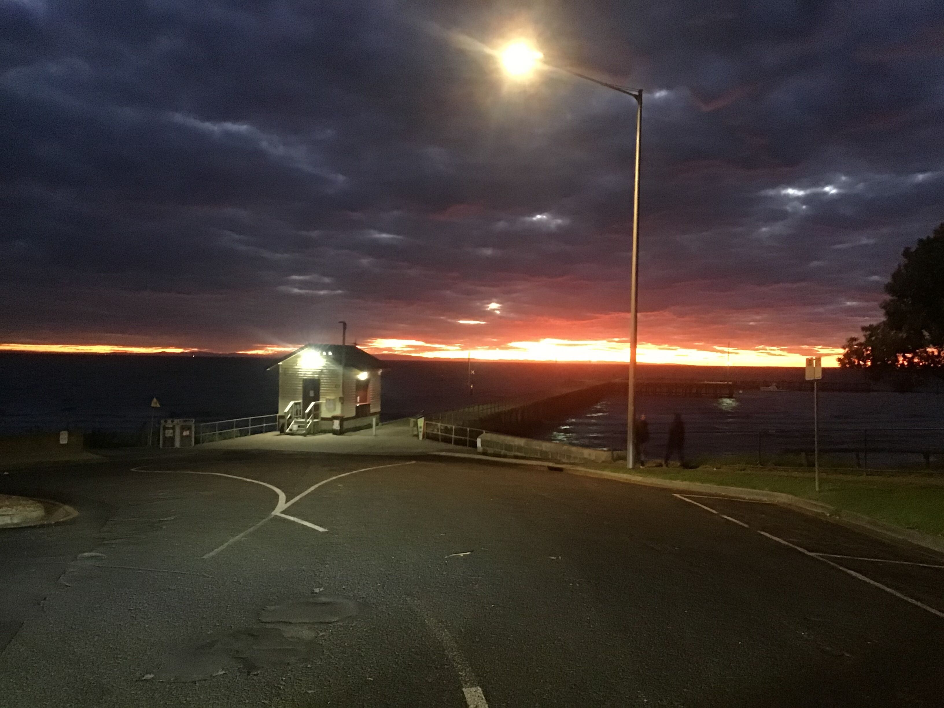 A coastal road leading to a small, illuminated lighthouse structure at sunset.