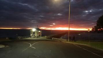 A coastal road leading to a small, illuminated lighthouse structure at sunset.