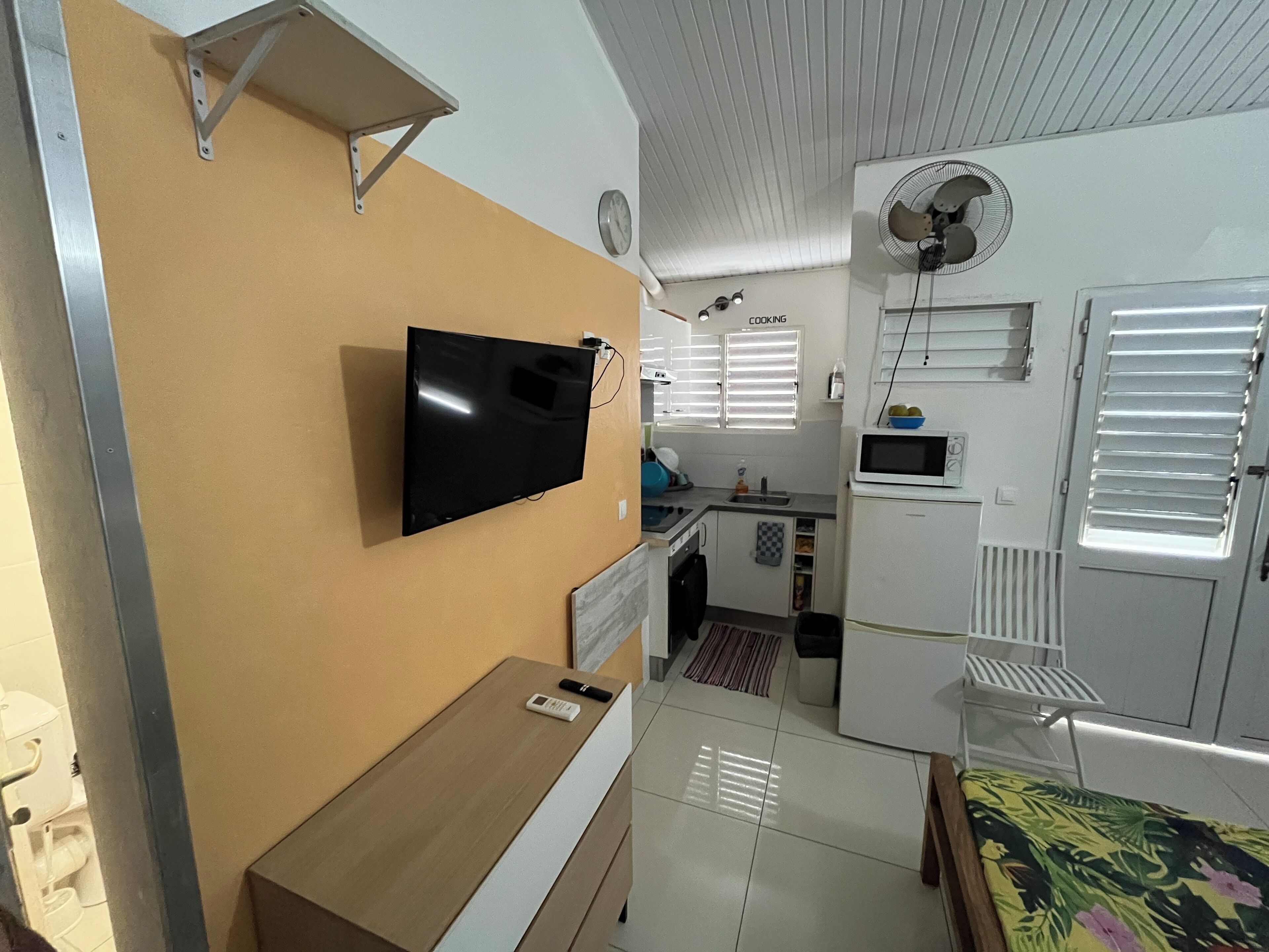 A compact living space with a flat-screen TV mounted on the wall, a small wooden table, a bed with a floral bedspread, a kitchenette with a microwave, and a ceiling fan.