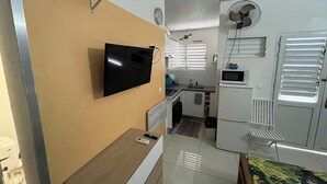 A compact living space with a flat-screen TV mounted on the wall, a small wooden table, a bed with a floral bedspread, a kitchenette with a microwave, and a ceiling fan. - Studio Vacances Martinique 3 ilets (Trois ilets)