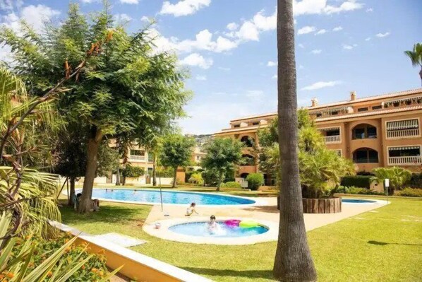 A pool area with multiple pools, surrounded by well-maintained grass and trees, and a building with a traditional architectural style in the background.
