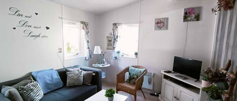 Un salon moderne avec un canapé bleu foncé, une table basse blanche, un fauteuil en osier, un téléviseur posé sur un meuble blanc et un mur orné d’une citation inspirante.