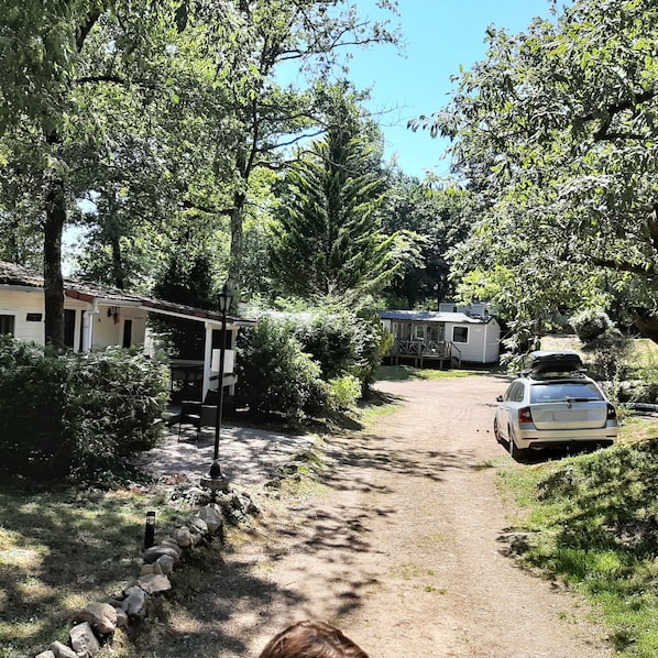Un chemin de gravier mène à un camping-car blanc garé sur la droite, entouré d’arbres et bordé d’un petit bâtiment sur la gauche.