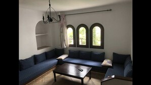 Una habitación con una mesa de centro de madera oscura, un sofá azul con cojines estampados y tres ventanas arqueadas que dejan entrar luz natural.