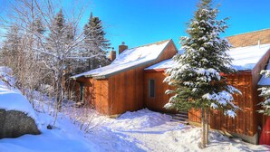 Eine Holzhütte mit schneebedecktem Dach, umgeben von Bäumen und blauem Himmel.