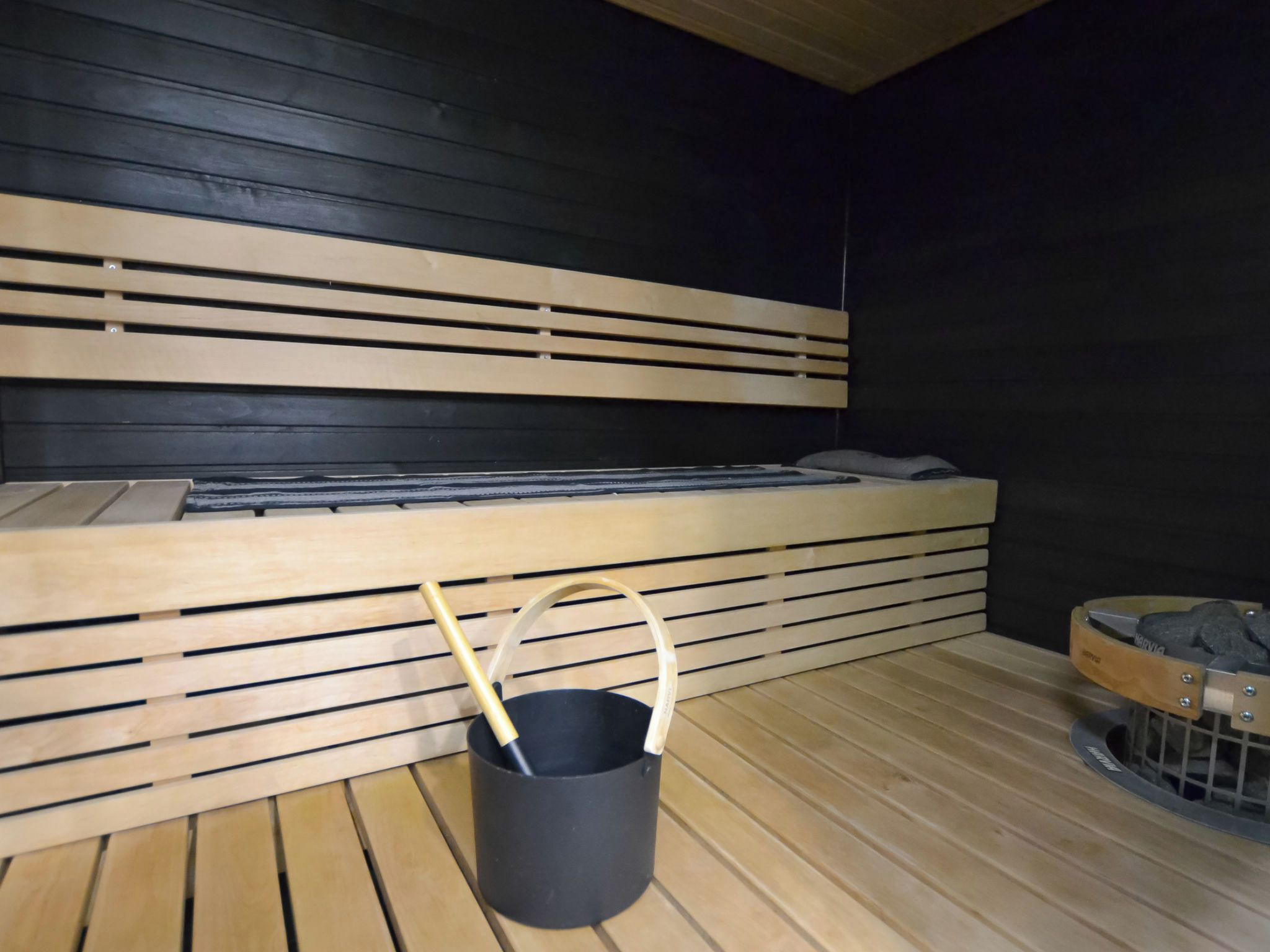 A sauna room with a wooden bench, a black bucket, and a stone container.