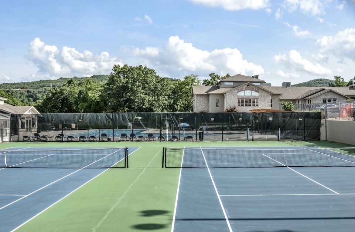 Ein Tennisplatz im Freien mit Netz, umgeben von einem Zaun, und ein Gebäude im Hintergrund.
