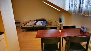 Una habitación pequeña y acogedora con piso de madera, una mesa roja y dos sillas negras. Tiene una cama con una manta y almohadas estampadas, una mesita con una lámpara y una ventana con cortinas decorativas.