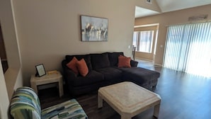 Ein Wohnzimmer mit einem Ecksofa, einem Couchtisch, einem gestreiften Hocker, einem gerahmten Bild und einem Wandbild.
