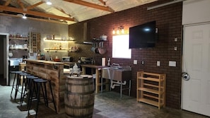 A rustic kitchen with a bar area, wooden stools, a large barrel, and a TV mounted on the brick wall.