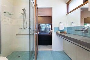 Casa de banho com cabine de duche em vidro, uma cama na divisão contígua e uma pia com espelho.