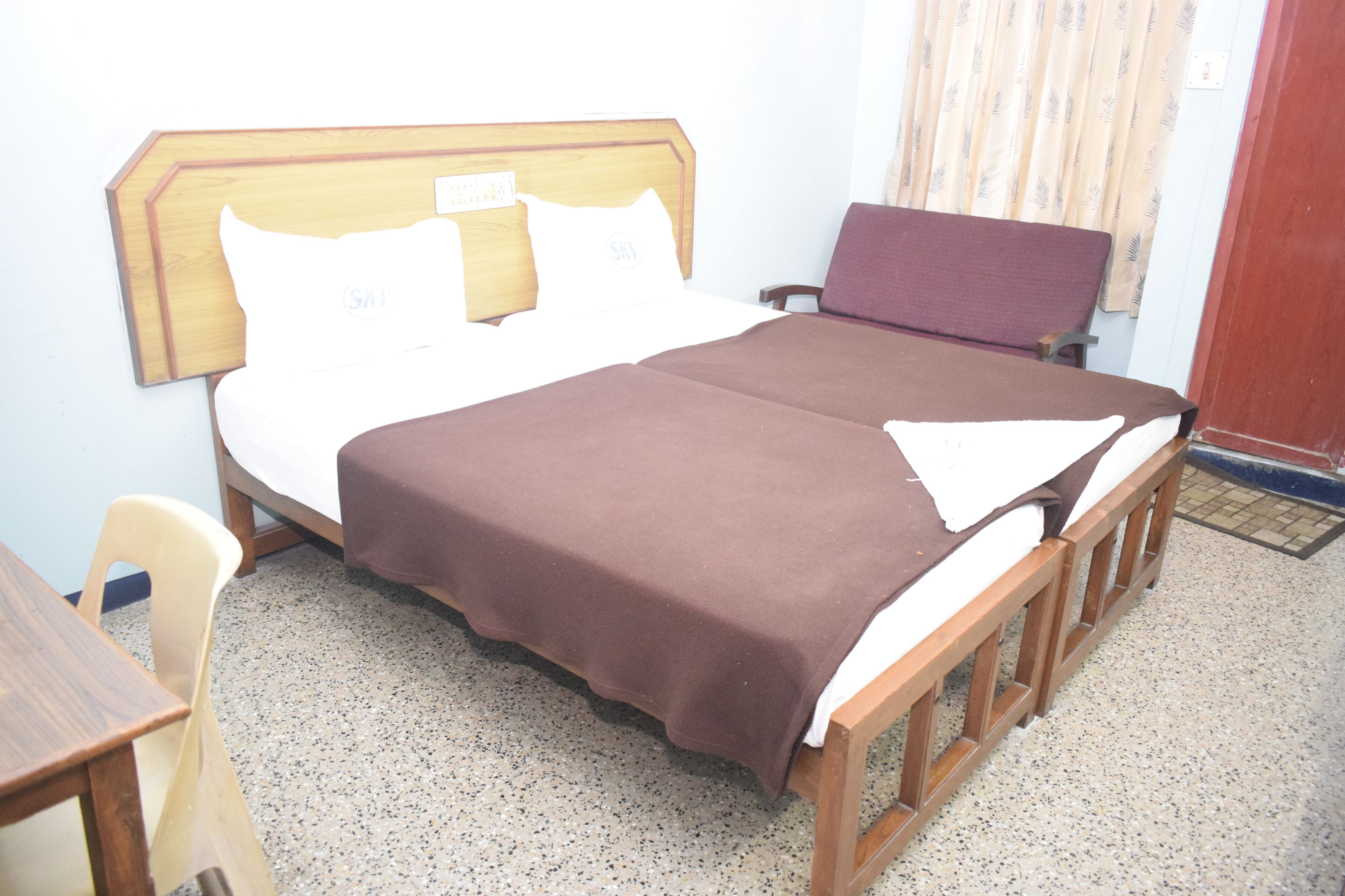 A single bed with a wooden headboard, a brown bedspread, and a white pillow with a logo. There is a wooden bench beside the bed and a plastic chair in the corner.