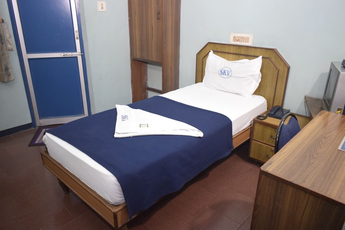 A single bed with a blue bedspread and white linens, a wooden nightstand with a telephone, a desk with a chair, and a wardrobe with a mirror.