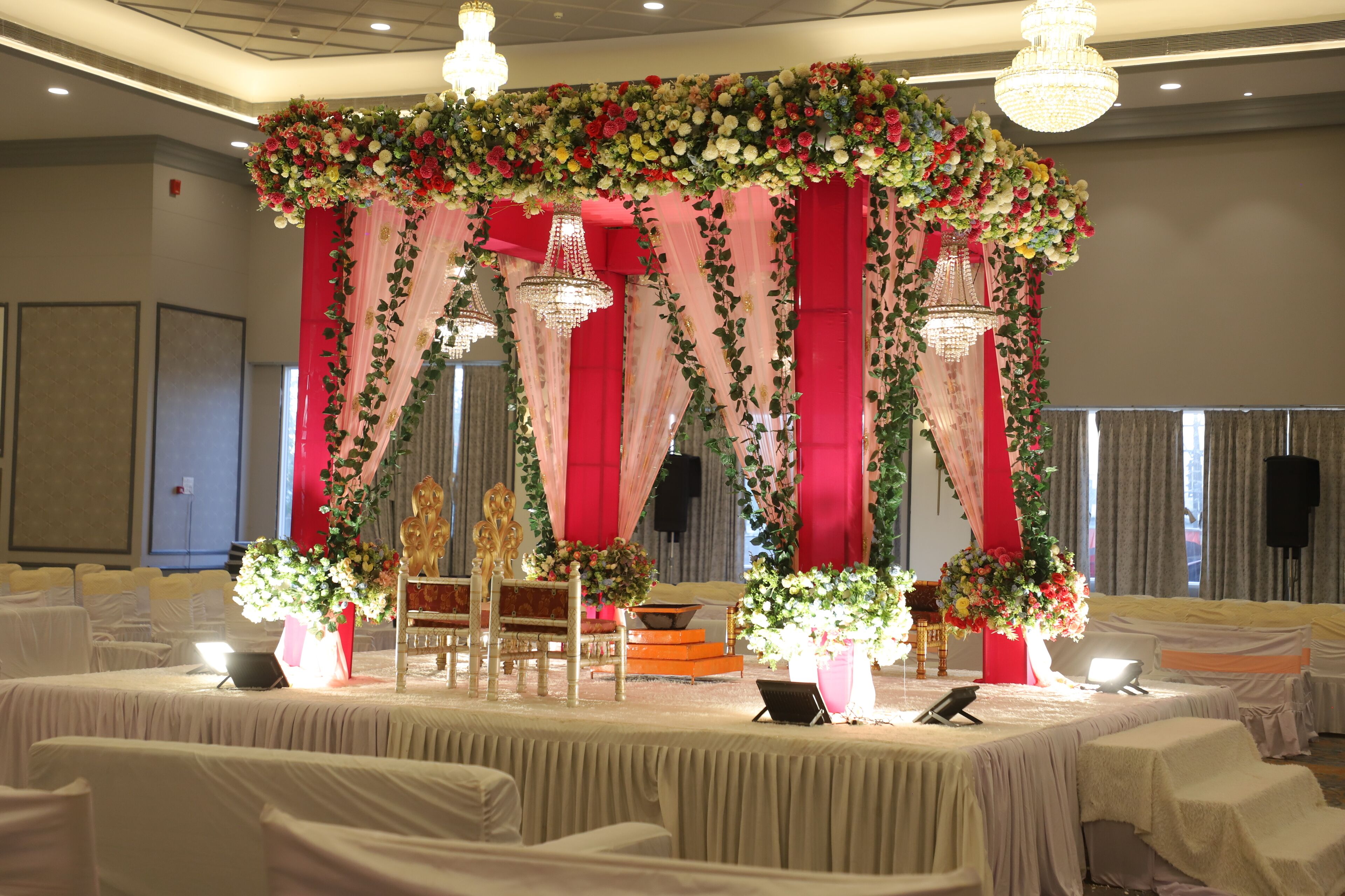 A banquet hall with round tables, chairs, and a stage with floral decorations and chandeliers.