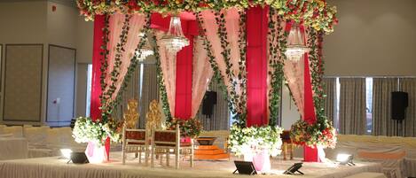 A banquet hall with round tables, chairs, and a stage with floral decorations and chandeliers.