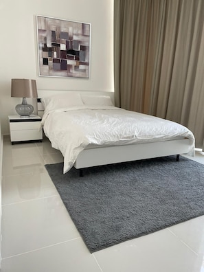 A modern bedroom with a white bed, a grey rug, a nightstand with a lamp, and a painting on the wall.
