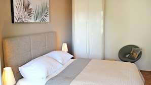 Een netjes opgemaakt bed met witte lakens en een grijs hoofdbord, een bedlampje en een groene stoel met kussen.