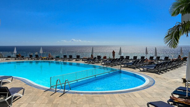 A poolside area with sun loungers and umbrellas, a clear view of the ocean, and a tiled deck.